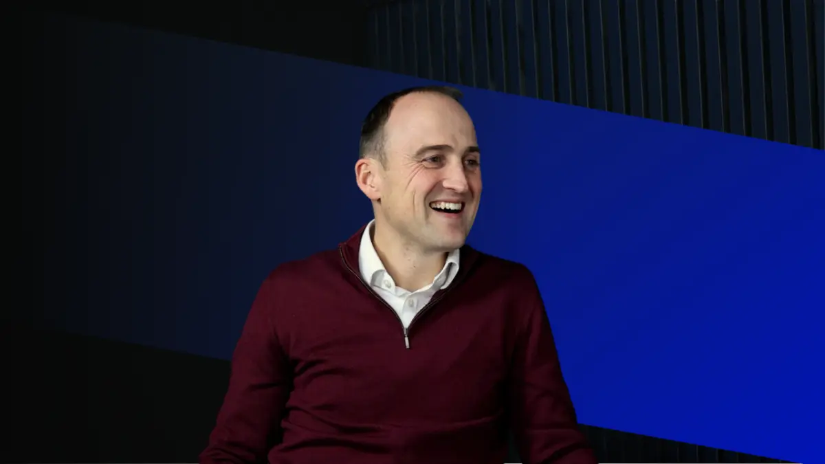 A man in a burgundy sweater smiling while sitting in front of a dark background with blue geometric shapes.