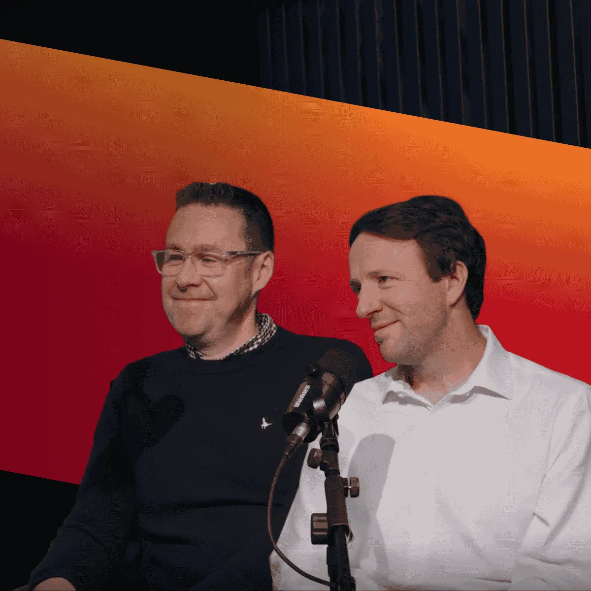 Two men stand beside a microphone on a stage, with a red-to-orange gradient backdrop behind them.