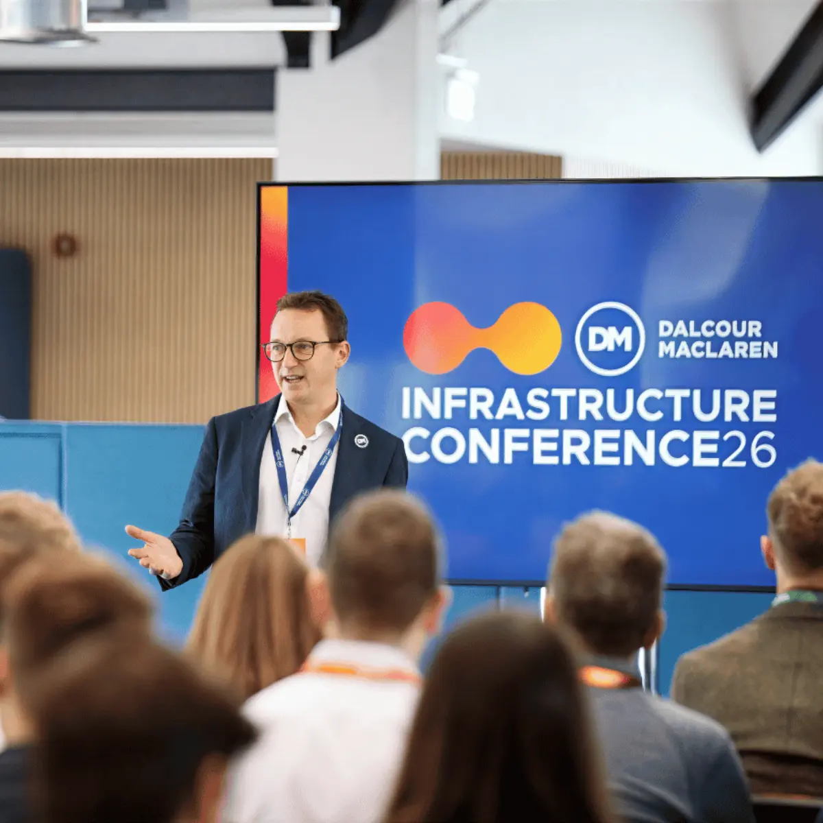 A man in glasses speaks to an audience in front of a screen reading Infrastructure Conference 26 and Dalcour Maclaren, with attendees seen from behind in a conference room.
