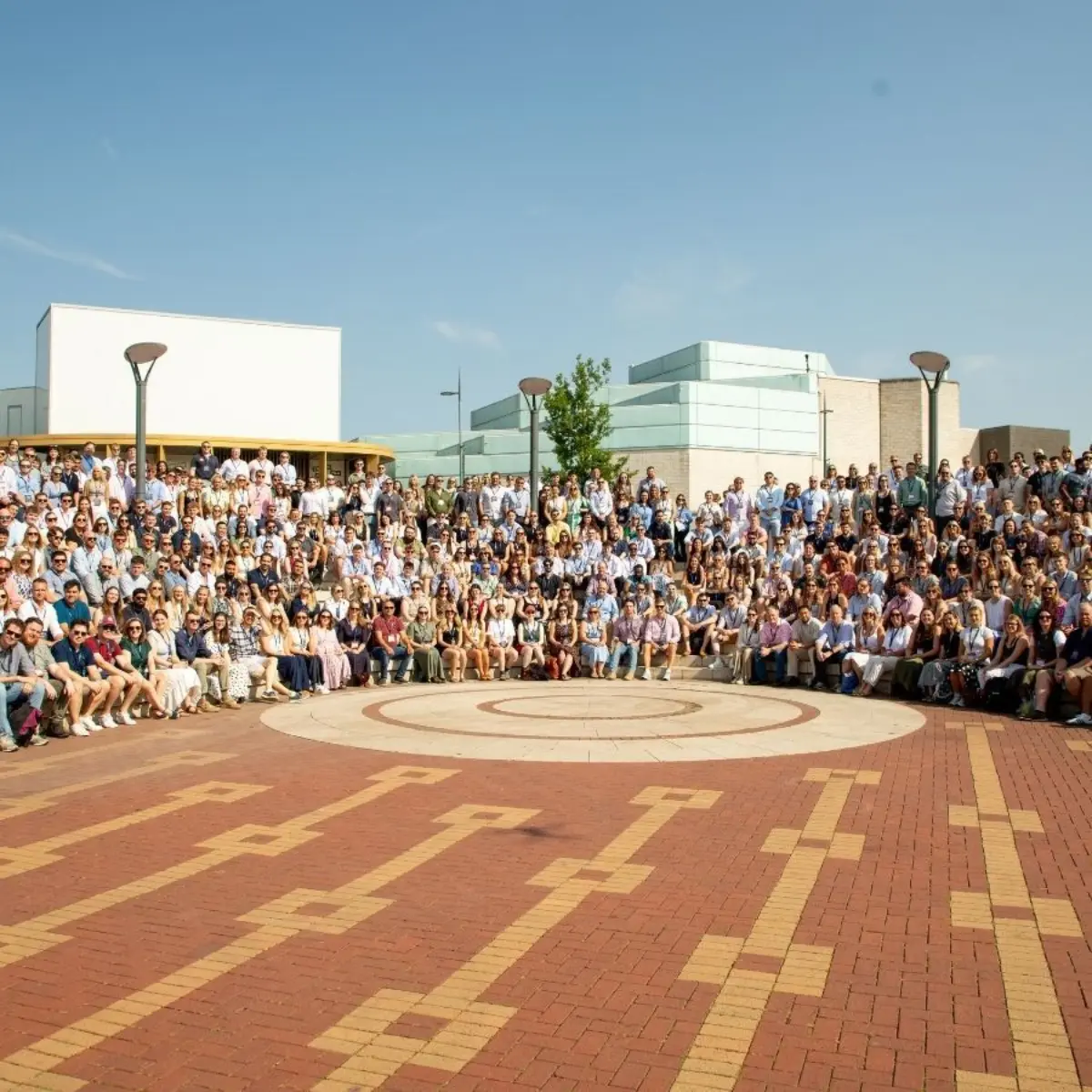 A large group of people gathered outdoors in front of modern buildings, seated and standing around a circular paved plaza.
