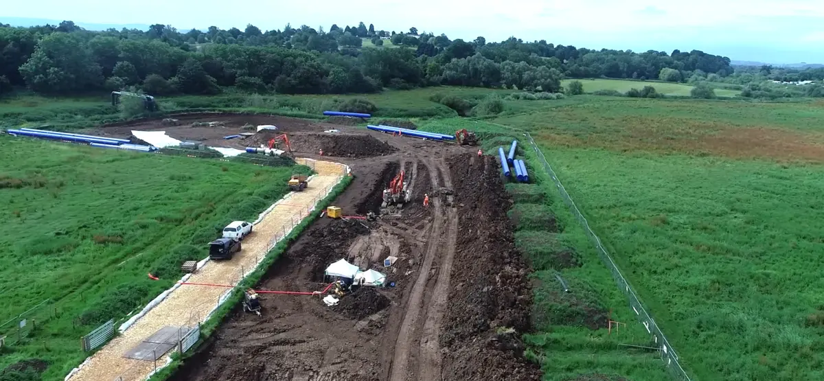 An aerial view of a construction site with heavy machinery and vehicles working on a dirt road. The area is surrounded by green fields and trees in the background under a cloudy sky.