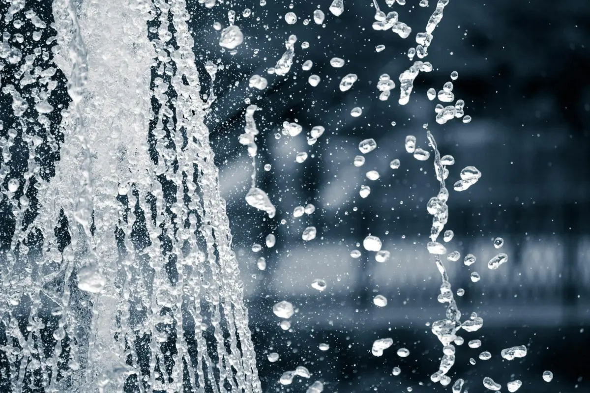 Close-up of water droplets splashing from a fountain against a blurred background. The image captures the motion and texture of the water as it cascades.