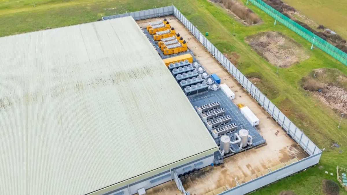 An aerial view of a large industrial building with a green metal roof. Adjacent to the building, various large equipment and generators are arranged on a concrete surface, surrounded by a security fence and grassy areas.