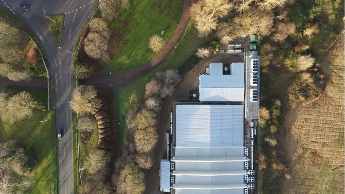 An aerial view of a large building with a flat roof and several ventilation units on top, surrounded by trees. Nearby, there are winding roads and patches of green grass, indicating a rural or suburban setting.