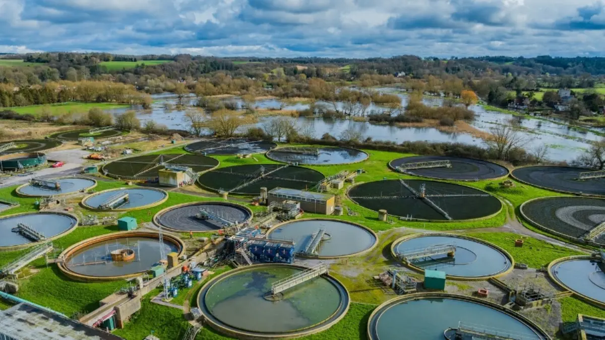 An aerial view of a wastewater treatment facility featuring multiple circular tanks surrounded by green grass. In the background, a river can be seen, along with trees and fields under a cloudy sky.