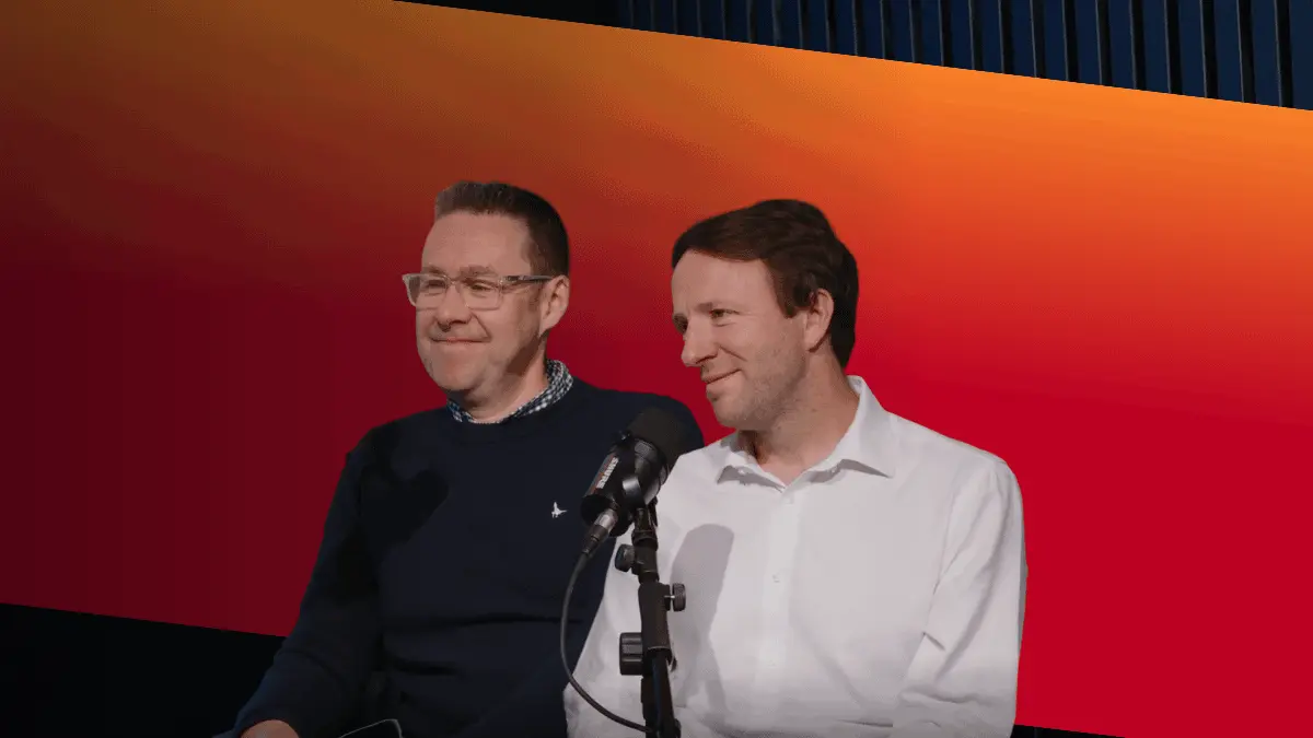 Two men stand beside a microphone in front of a red and orange gradient screen, with one wearing a navy sweater and the other a white shirt.