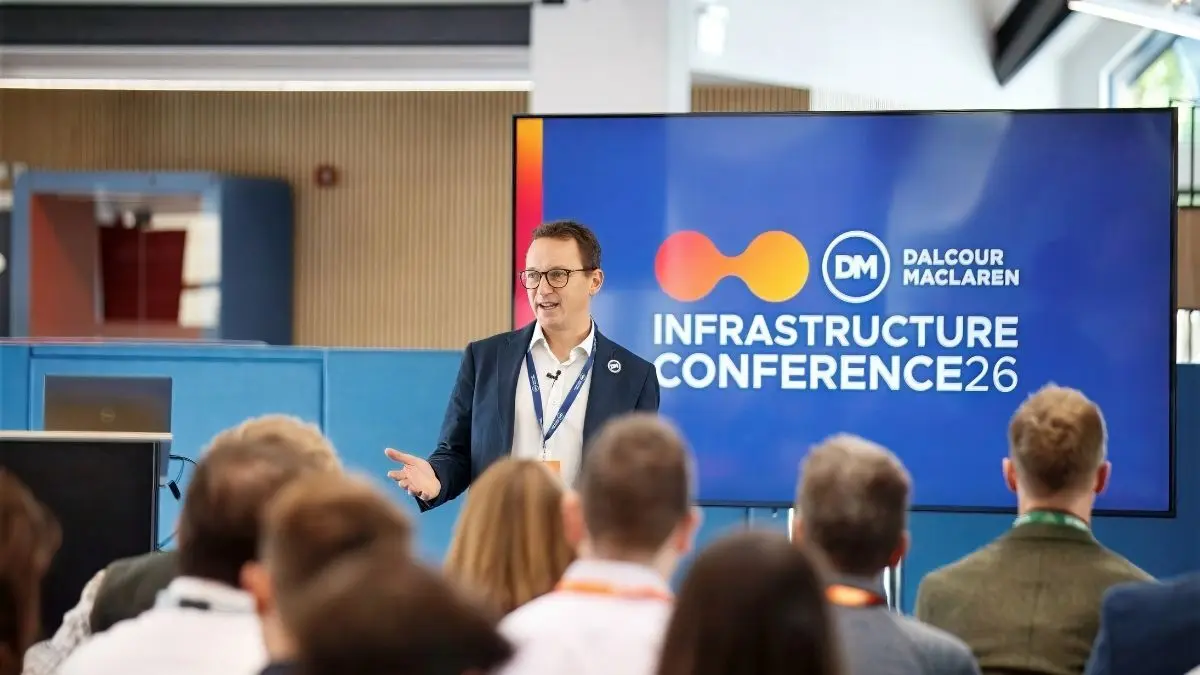 A man in a blazer speaks at the Dalcour Maclaren Infrastructure Conference 26, with an audience seated in the foreground and a presentation screen behind him.