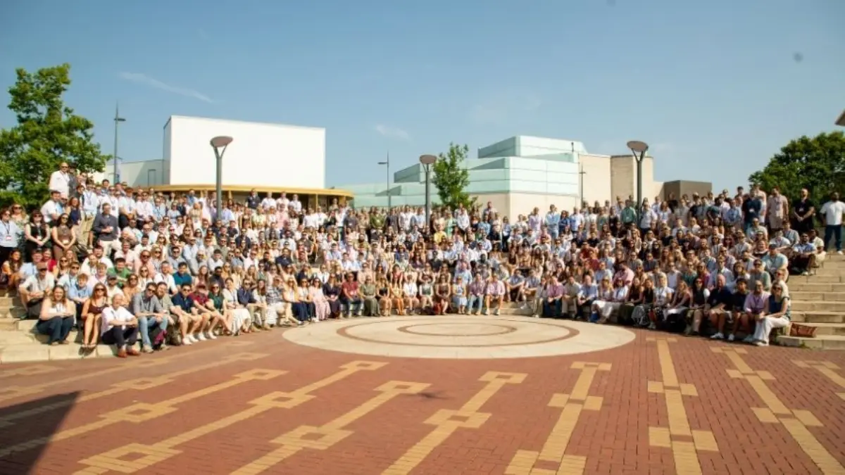 A large group of people poses outdoors on a brick plaza with a circular pattern, in front of modern campus buildings and trees.