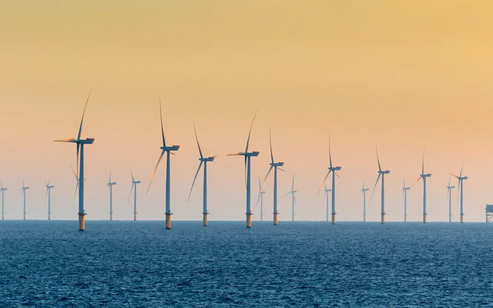 A series of wind turbines lined up in the ocean, with the sun setting in the background, creating a gradient of colors in the sky. The water is calm, reflecting the turbines.