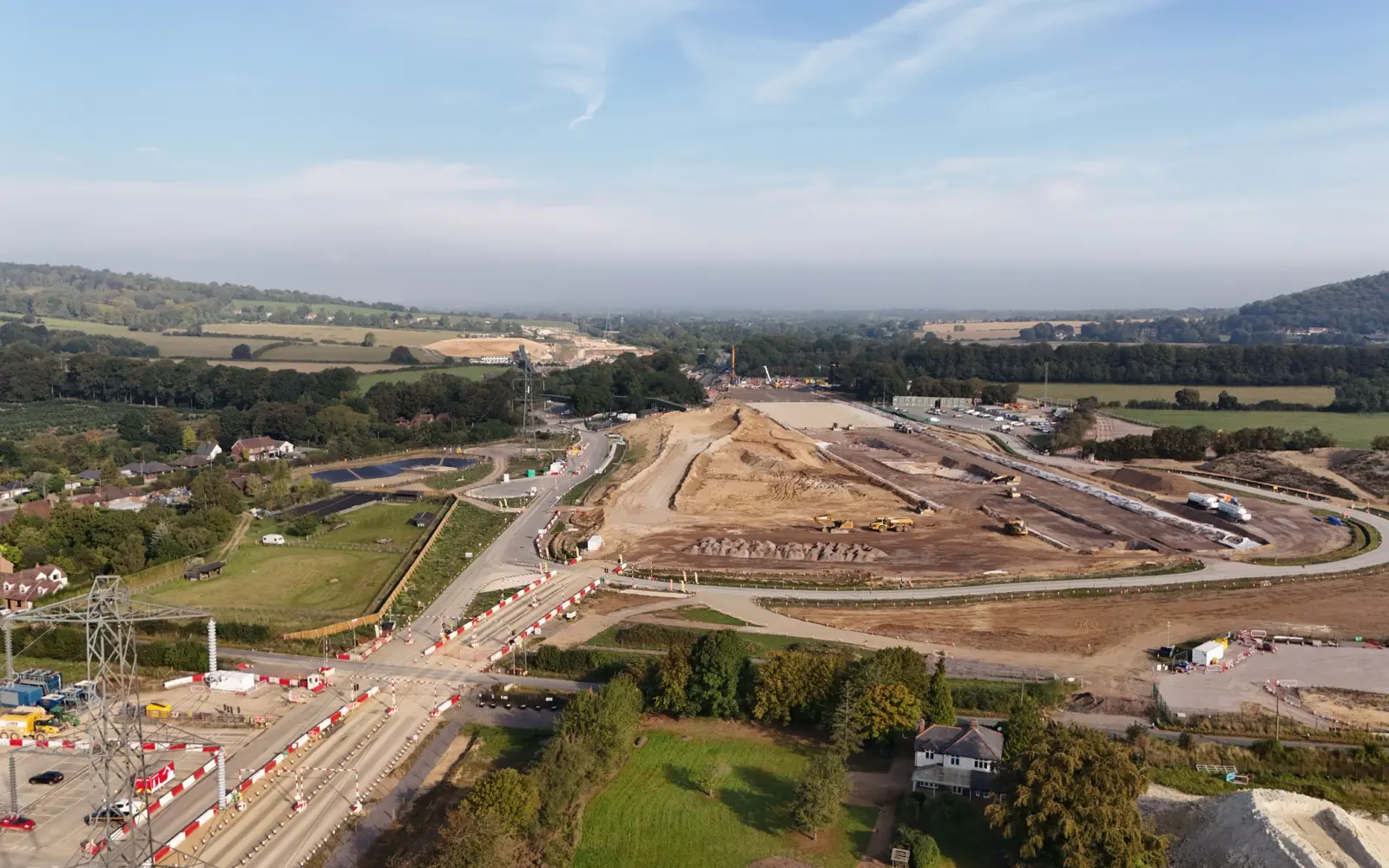 Aerial view of a construction site in a rural area, featuring large earth-moving equipment and cleared land. Surrounding fields and a distant wooded area are visible, with roads and traffic cones indicating ongoing work in the vicinity.