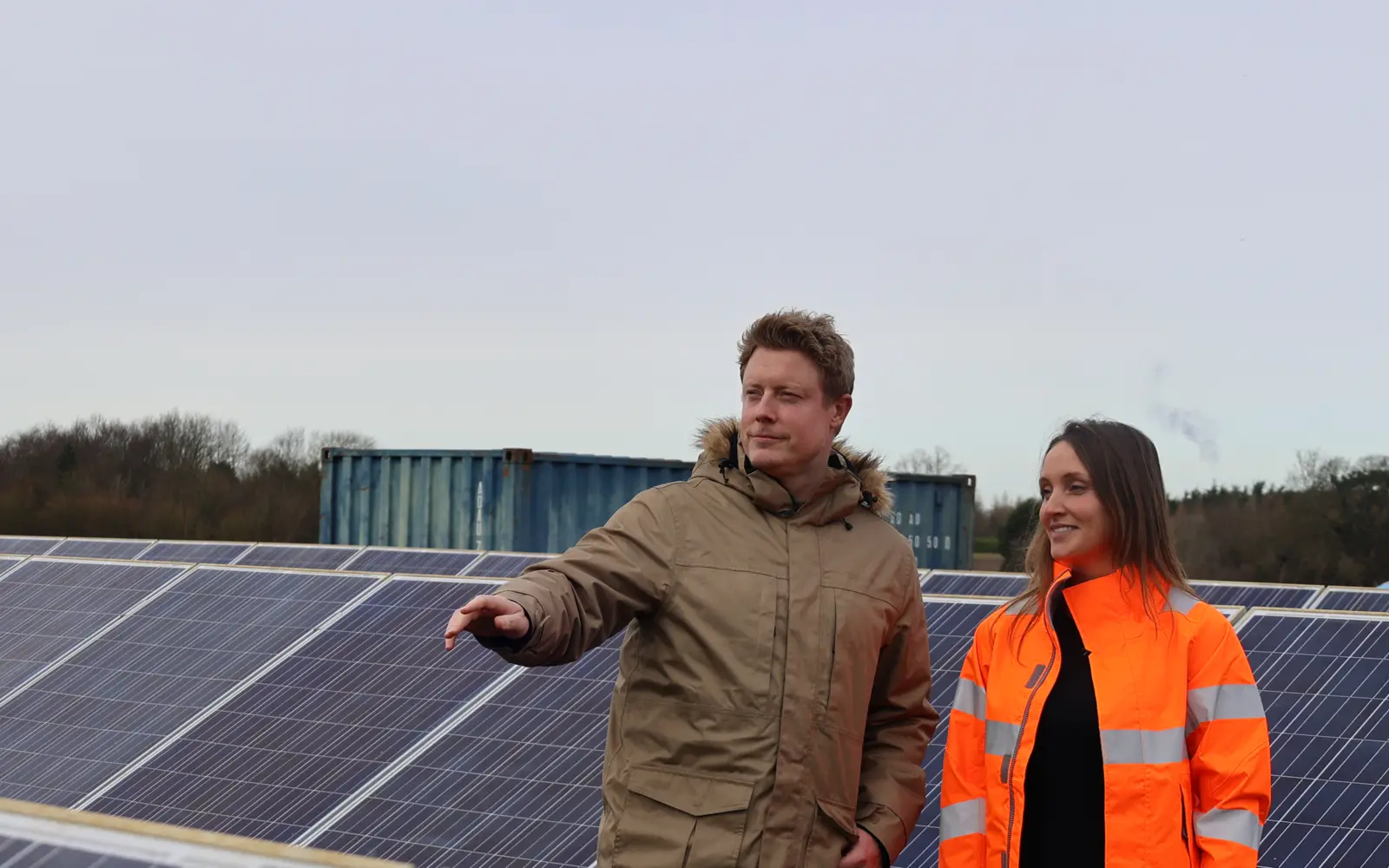 Two individuals, one in a tan coat and the other in an orange safety jacket, standing near solar panels with a cloudy sky in the background. They appear to be discussing something while looking out at the panels.