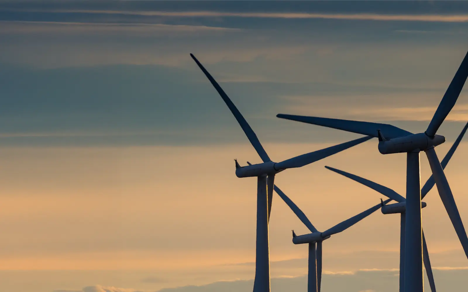 Silhouettes of wind turbines against a colorful sky during sunset, with soft clouds in the background.