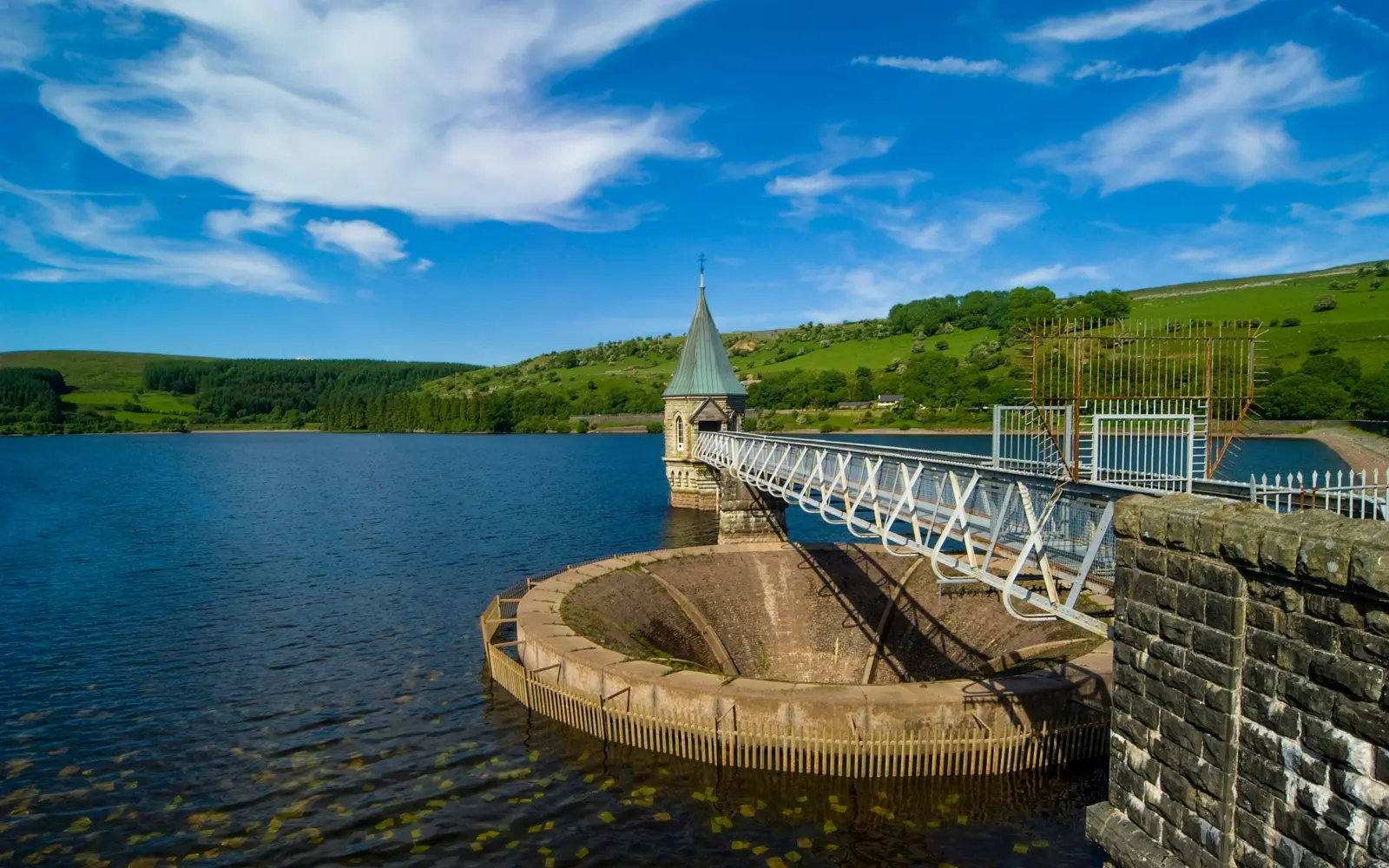 A scenic view of a reservoir with a stone tower and a bridge extending across the water. Lush green hills are visible in the background, while fluffy clouds float in a blue sky.