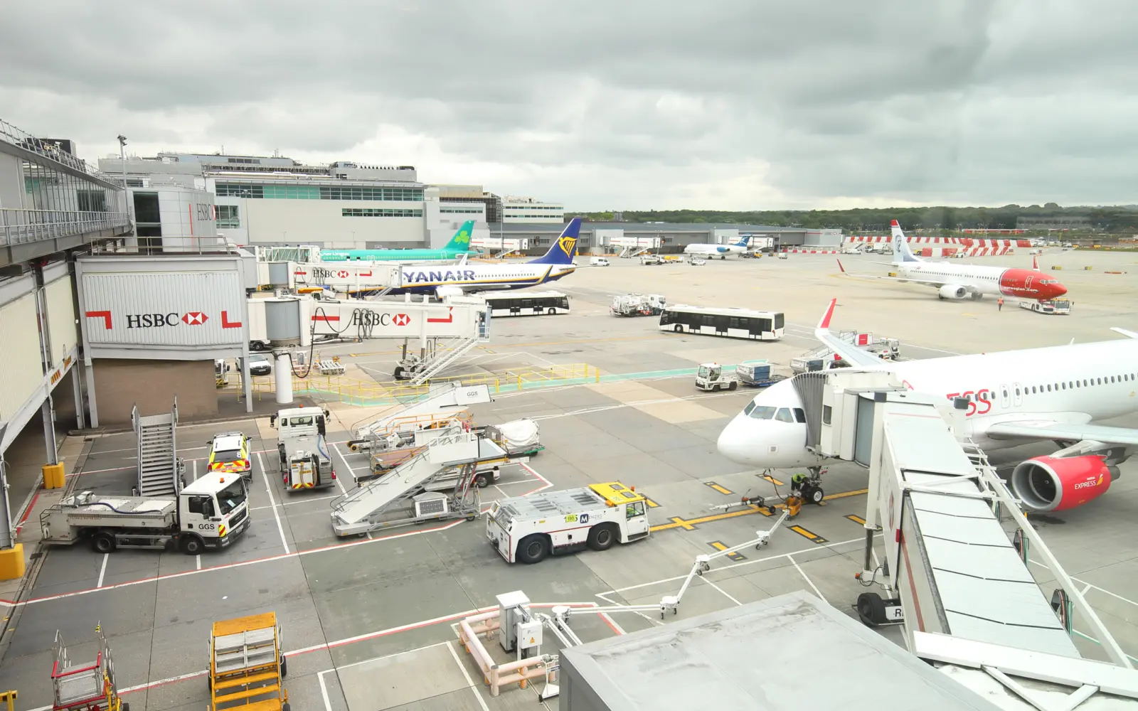 An airport scene showing multiple airplanes parked at gates, including a Yanair flight, and ground service vehicles assisting with luggage at a busy terminal.