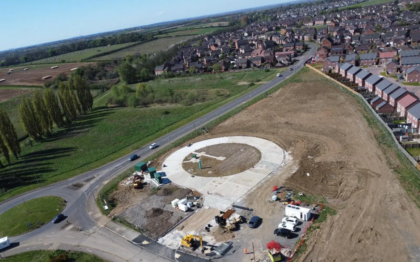 An aerial view of a construction site featuring a circular concrete foundation for a roundabout, surrounded by cleared land and vehicles. In the background, residential houses are visible alongside green fields.