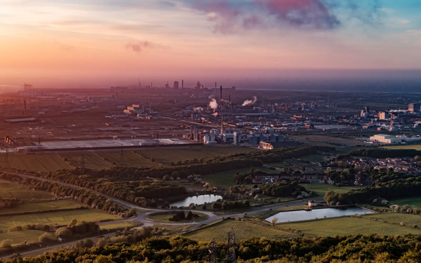 A panoramic view of an industrial landscape during sunset, featuring factories and smokestacks in the background, surrounded by green fields and bodies of water.