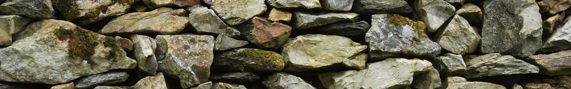 A close-up view of a dry-stone wall made up of various sizes and shapes of gray and tan stones, with patches of moss and grass visible at the base.