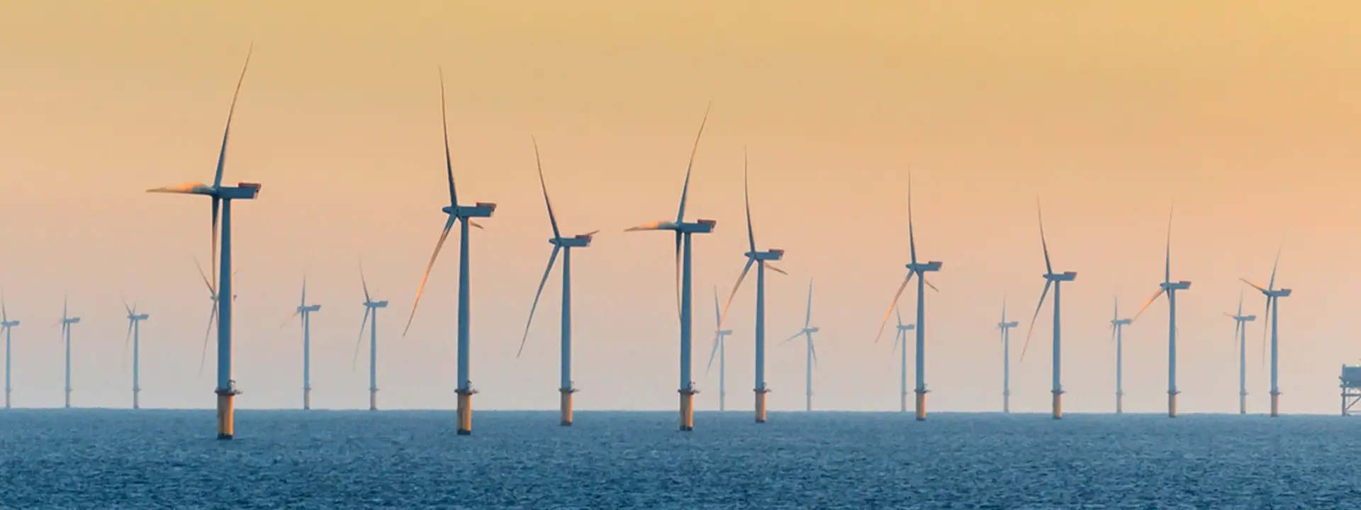 A series of wind turbines lined up in the ocean, with the sun setting in the background, creating a gradient of colors in the sky. The water is calm, reflecting the turbines.