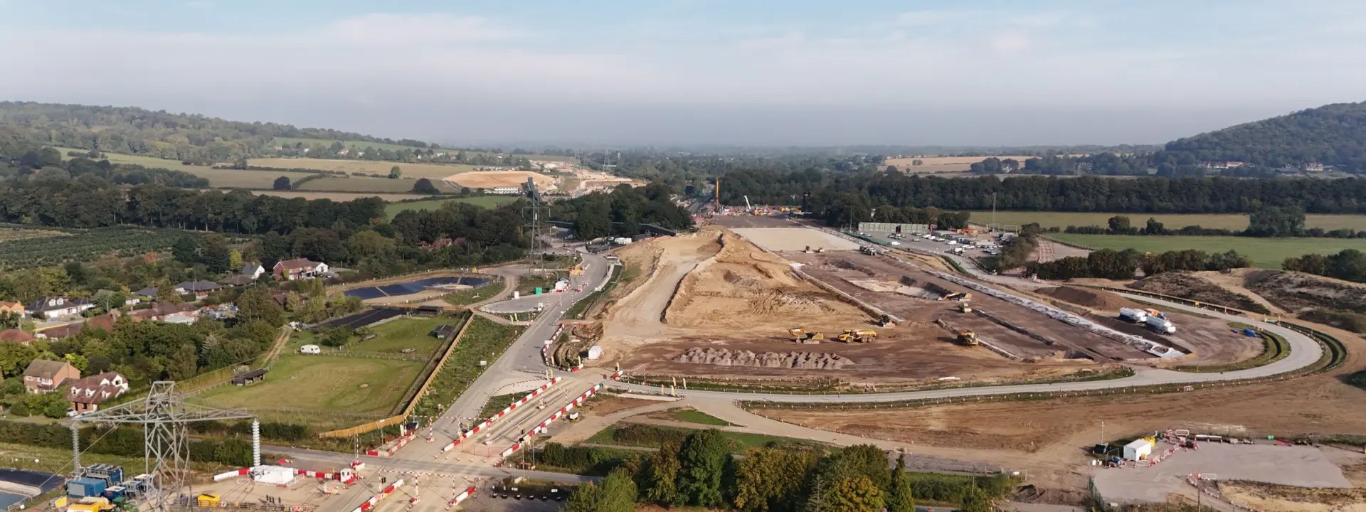 Aerial view of a construction site in a rural area, featuring large earth-moving equipment and cleared land. Surrounding fields and a distant wooded area are visible, with roads and traffic cones indicating ongoing work in the vicinity.