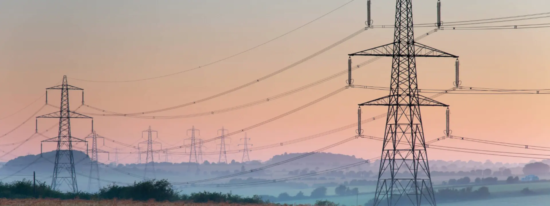 Multiple electricity pylons in a rural landscape at sunrise, with fog covering the fields and distant hills under a soft pastel sky.