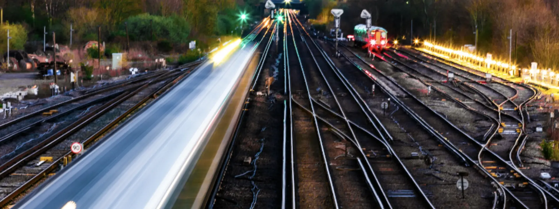 A view of railway tracks at dusk, with train lights creating streaks of light. Multiple tracks curve away into the distance, surrounded by trees and signaling lights.