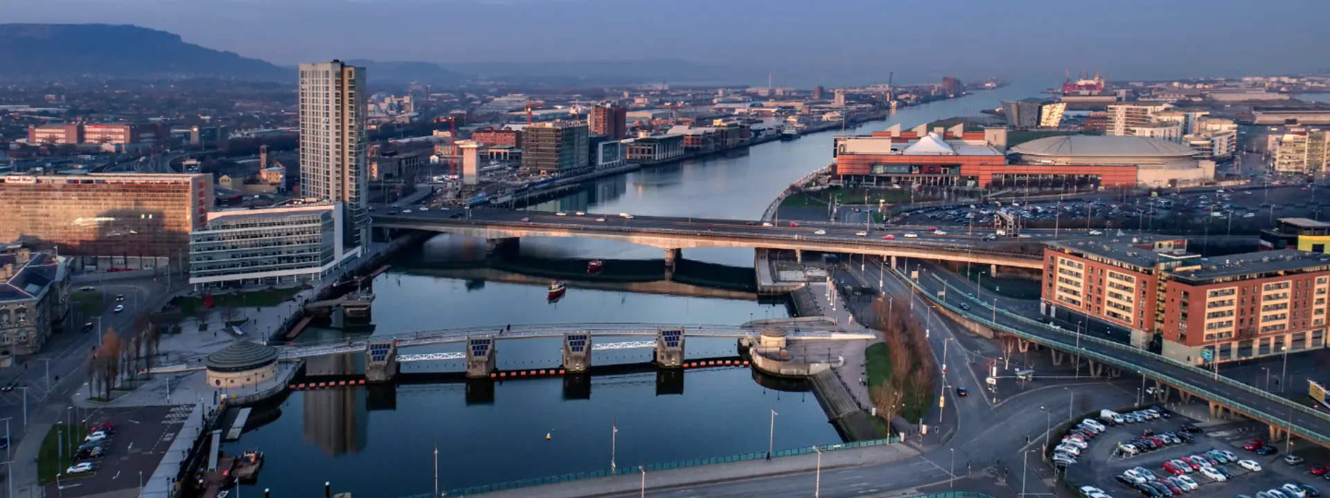 Aerial view of an urban area featuring a wide river, modern buildings, and bridges. A marina and parking lot are visible, along with cityscape in the background. The scene is bathed in early morning light.