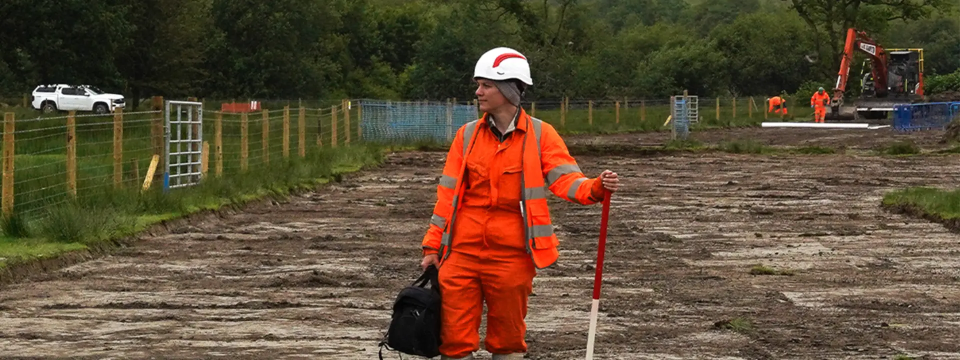 A construction worker in an orange safety suit and helmet stands on a cleared site, holding a measuring stick. Equipment and workers in the background indicate ongoing construction work.