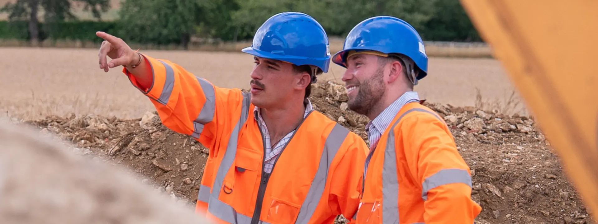 Two construction workers in orange safety vests and blue hard hats discussing a project. One is pointing towards the distance while the other listens attentively, with a rural landscape in the background.