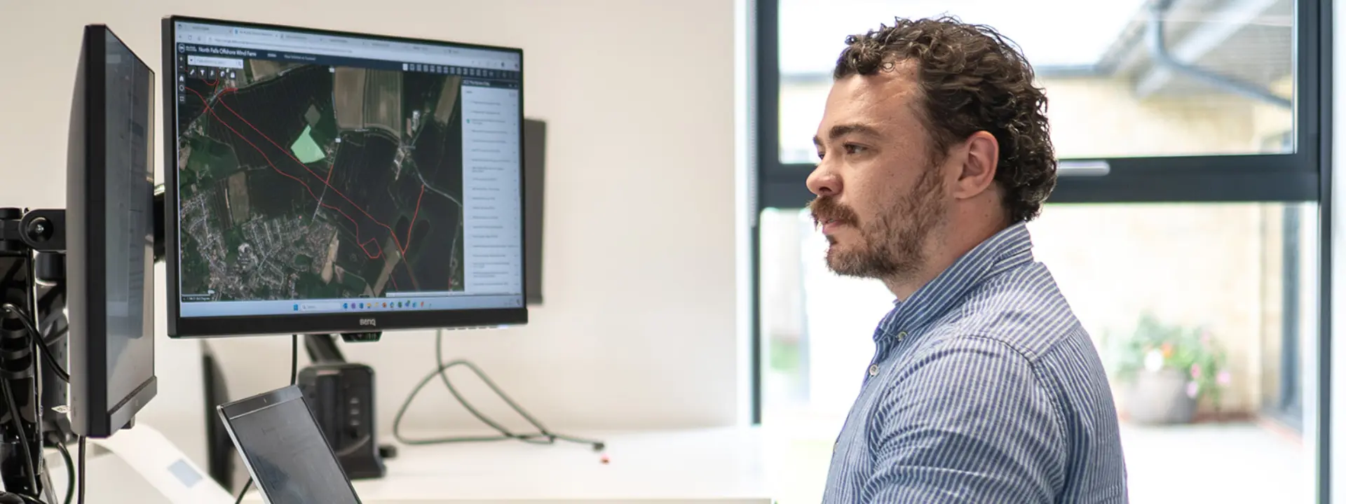 A man sitting at a desk working on a computer, focused on a large monitor displaying a digital map. The workspace is bright with natural light coming from a window.