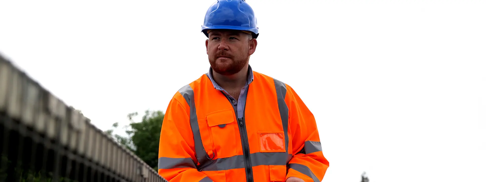 A man wearing an orange safety jacket and a blue hard hat stands near a construction site, holding a tablet. The sky is overcast with trees visible in the background.