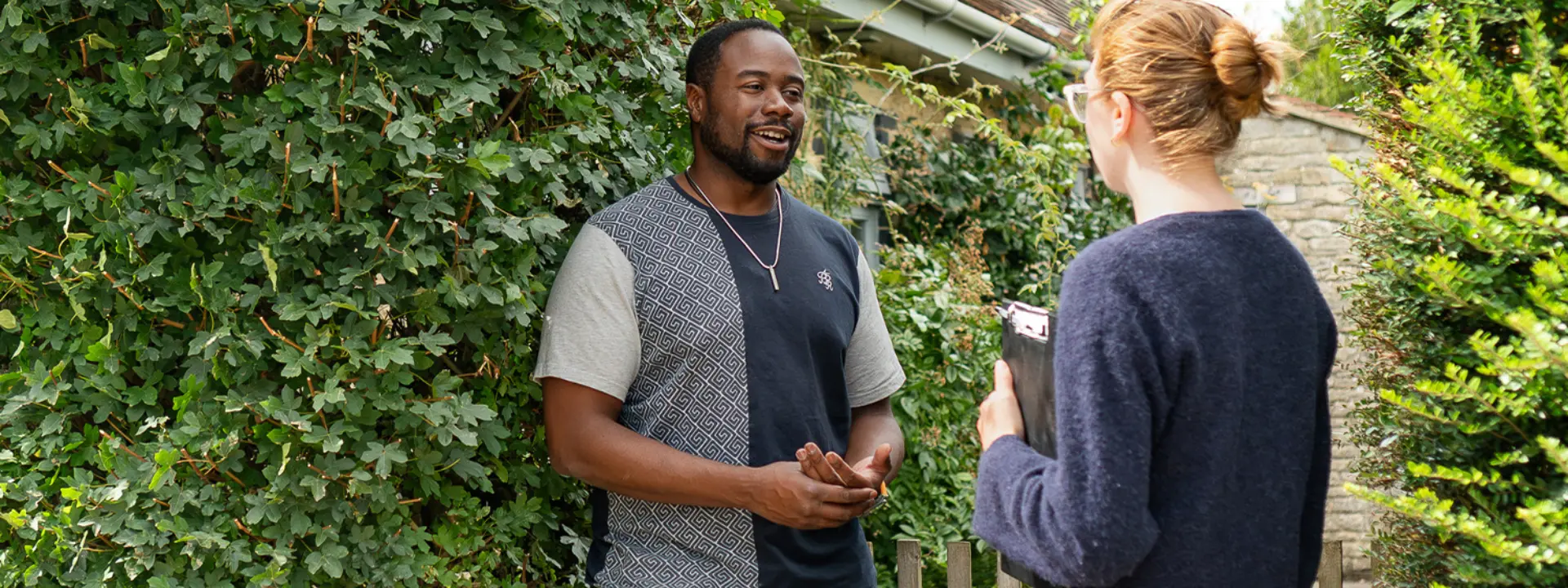 A man in a casual t-shirt is smiling and engaging in conversation with a woman holding a clipboard in front of a house covered in greenery.