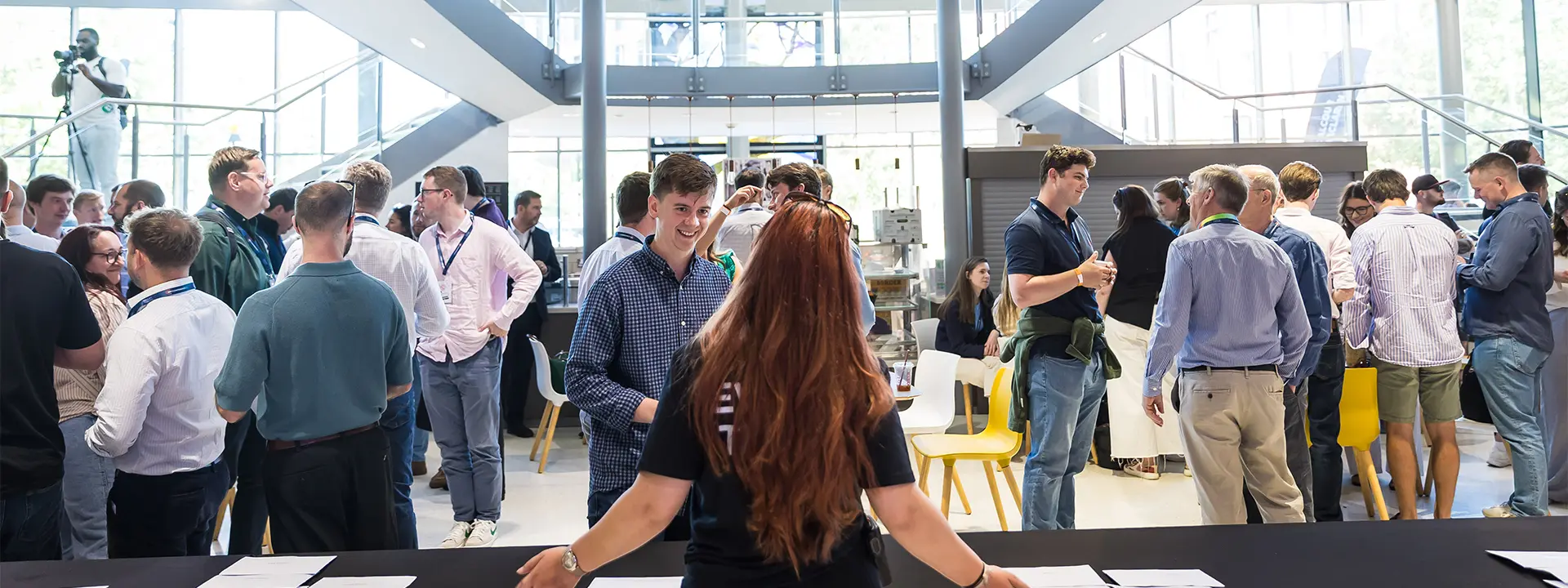 A group of young professionals networking in a modern atrium, with a woman in a black shirt welcoming attendees at a registration table. Bright windows and a staircase are visible in the background.