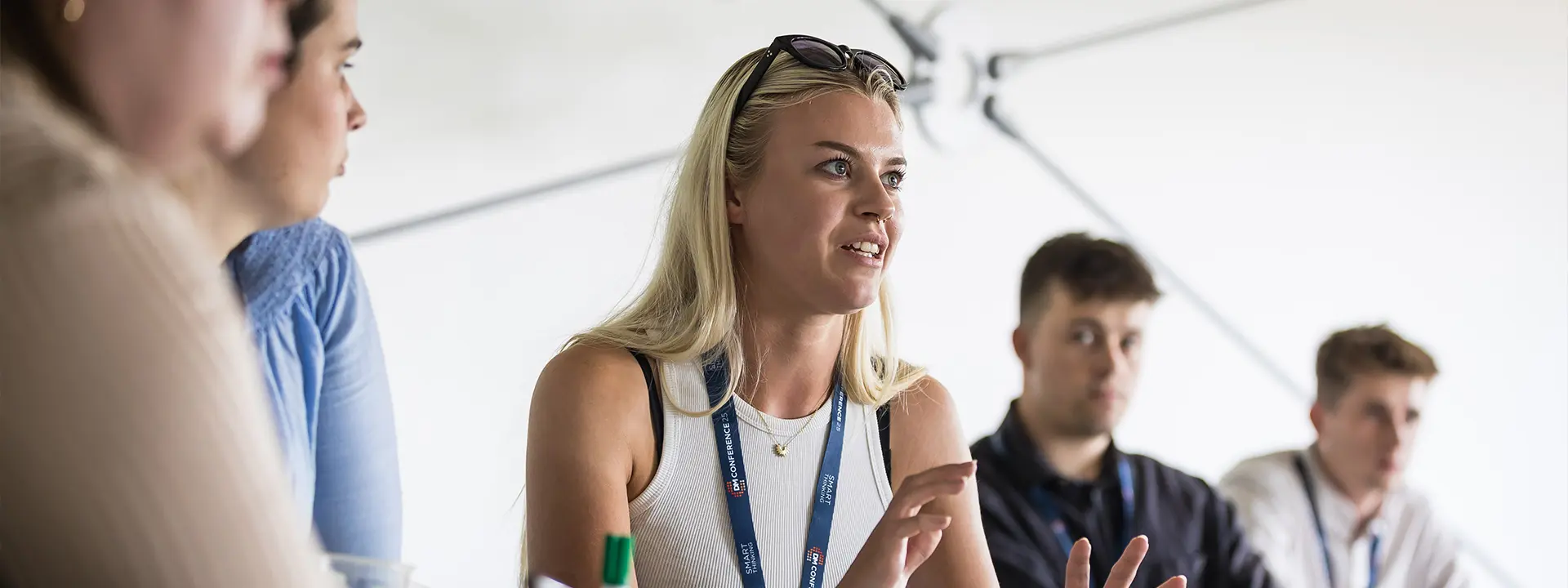 A group of individuals engaged in a discussion around a table, with a woman in the foreground speaking. She has long blonde hair and is wearing a sleeveless top. Others are listening attentively.