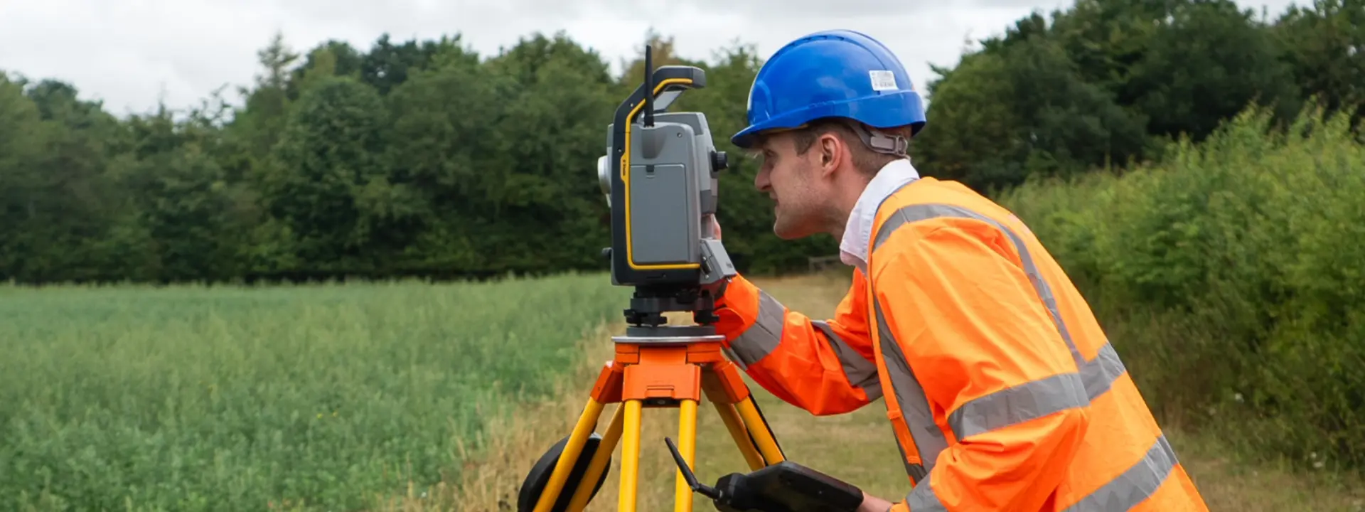 A construction worker in an orange safety jacket and blue hard hat uses a surveying instrument on a tripod in a grassy field, with trees in the background.