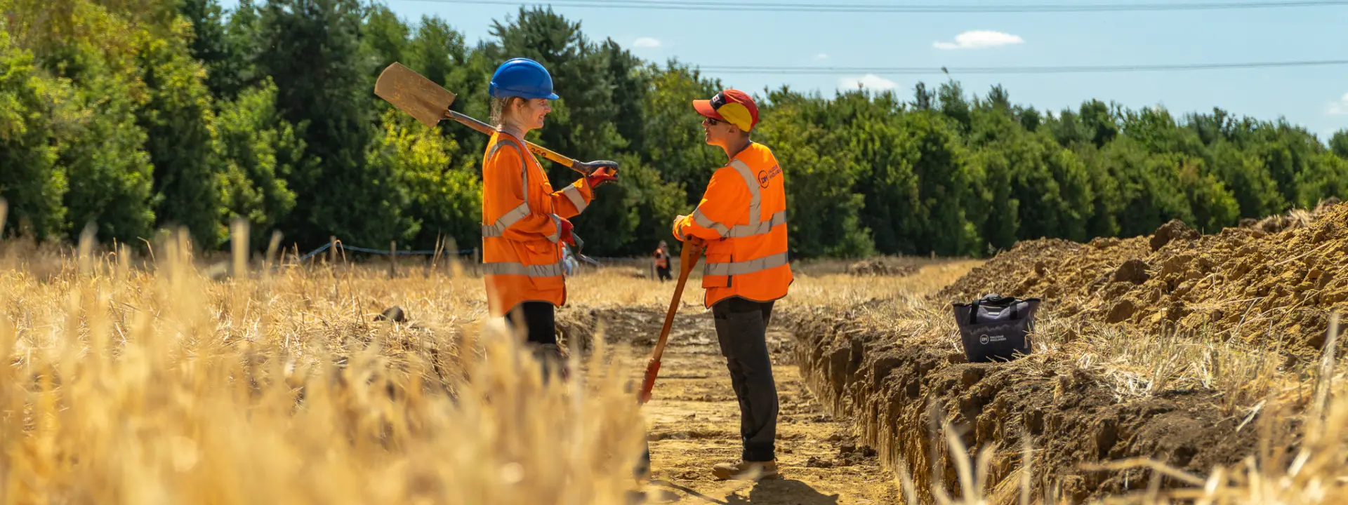 Two construction workers, wearing orange vests and helmets, are discussing near a trench in a field. In the background, trees and power lines are visible under a clear blue sky.