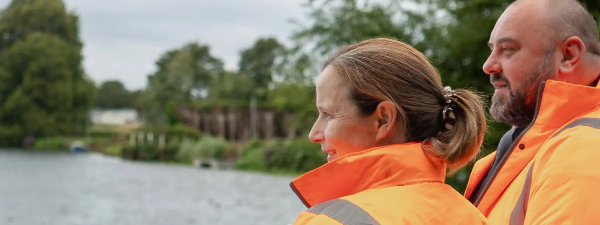 Two individuals wearing bright orange safety jackets are standing by a river, looking at the water. The background features greenery and a cloudy sky, creating a serene outdoor setting.