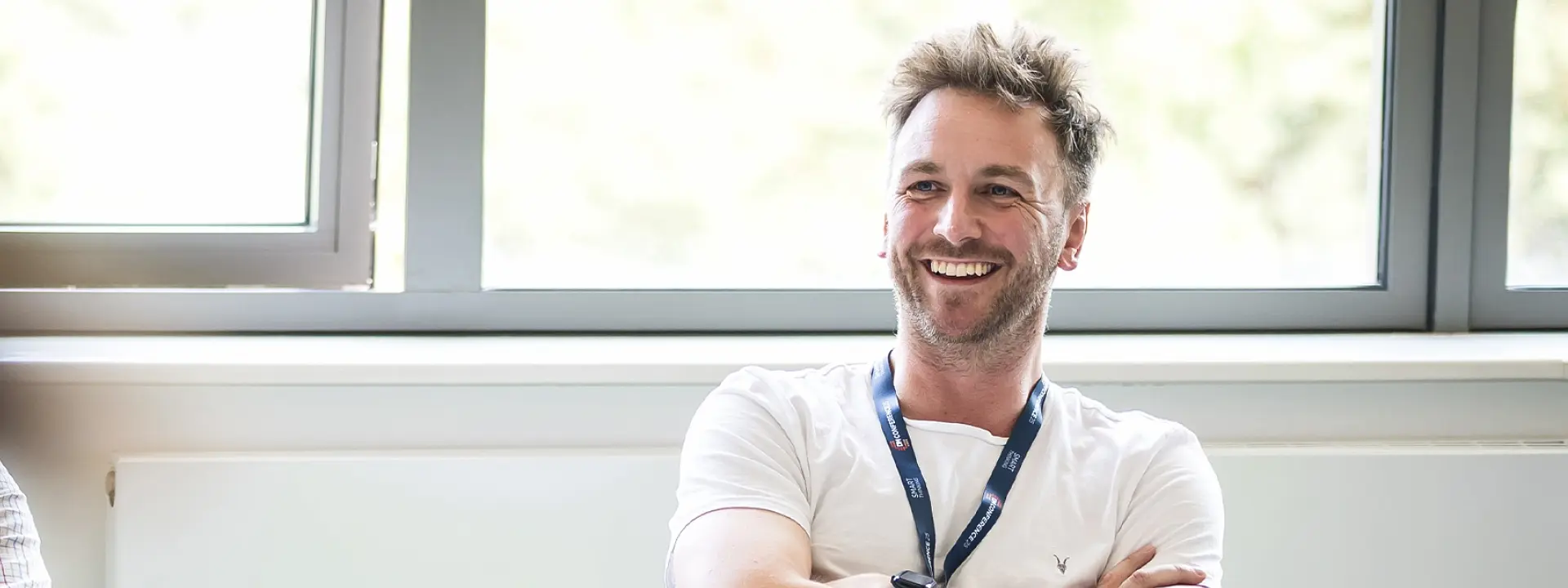 A man with short, light hair wearing a white t-shirt and a lanyard smiles while sitting in a bright room with large windows. The focus is on him, with some blurred figures in the foreground.