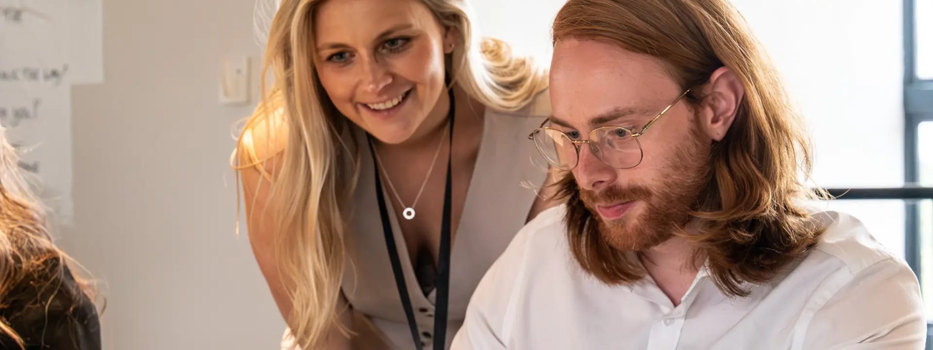 A woman standing beside a man seated at a laptop, both engaged in a discussion. The woman has long blonde hair and is wearing a light-colored dress. The man has long reddish-brown hair and glasses, focused on the screen.