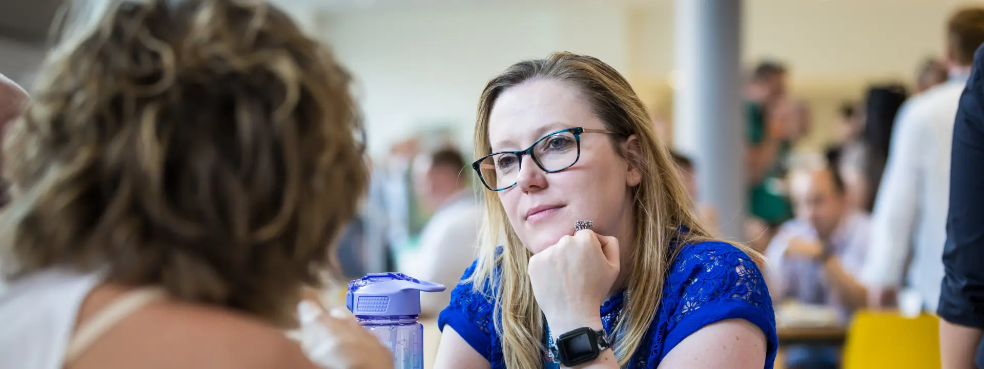 A woman with glasses wearing a blue shirt listens attentively while resting her chin on her hand at a table in a busy café, engaged in conversation with another person out of view.