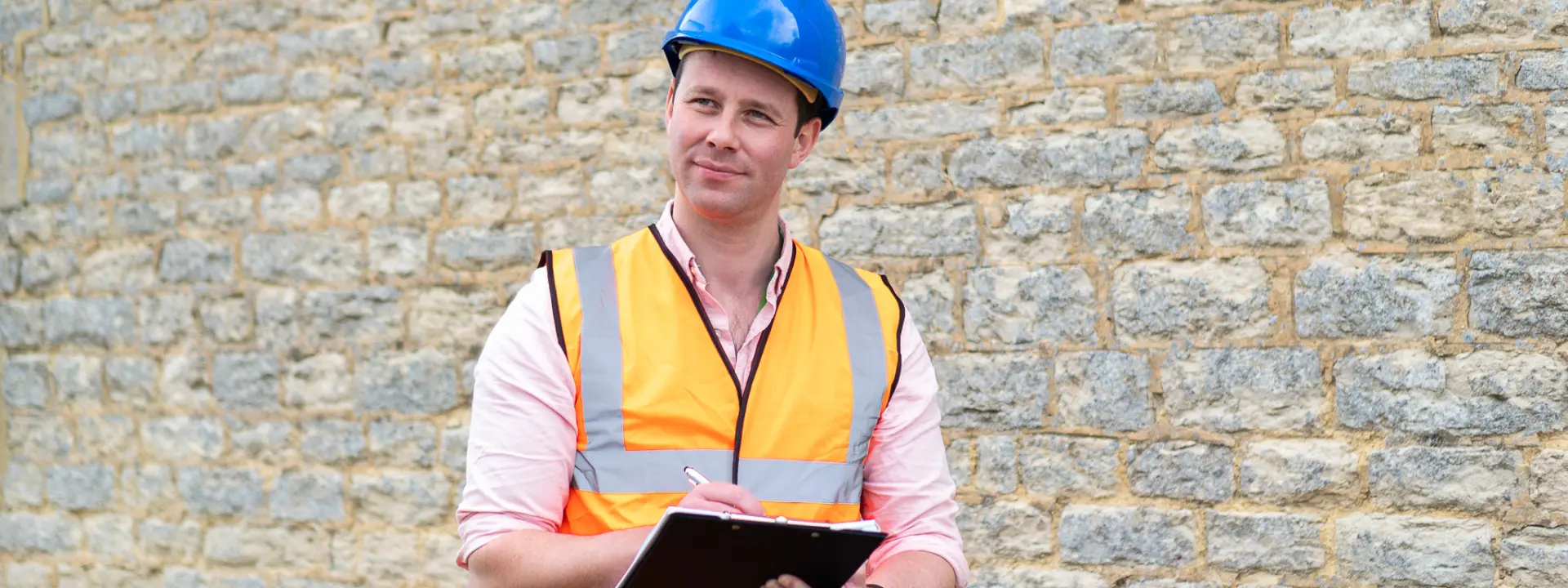 A construction worker wearing a blue hard hat and orange safety vest, holding a clipboard while standing in front of a stone wall.