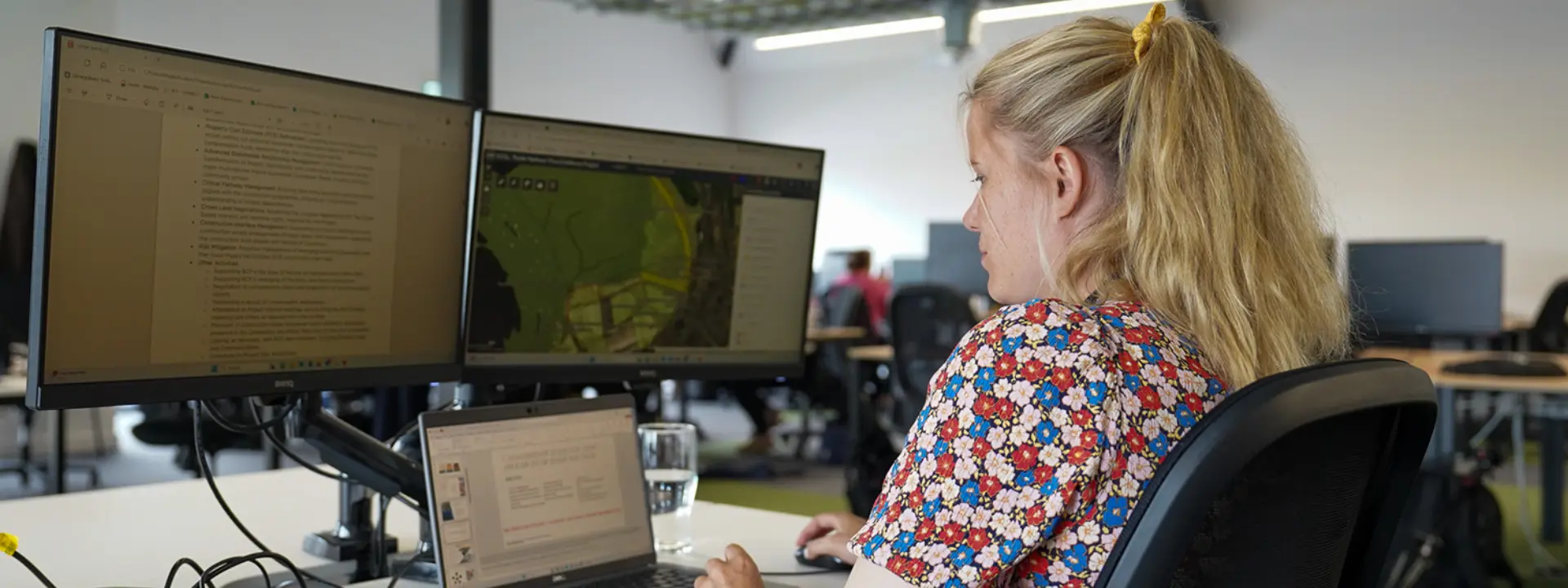 A woman with blonde hair sits at a desk in an office, using a laptop and two external monitors. The screens display text and a map. A glass of water is on the desk.