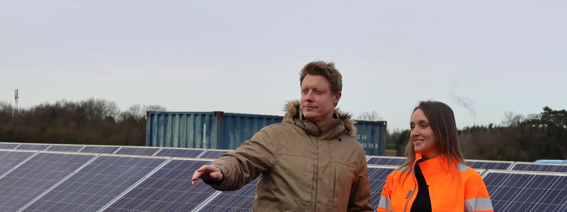 Two individuals, one in a tan coat and the other in an orange safety jacket, standing near solar panels with a cloudy sky in the background. They appear to be discussing something while looking out at the panels.