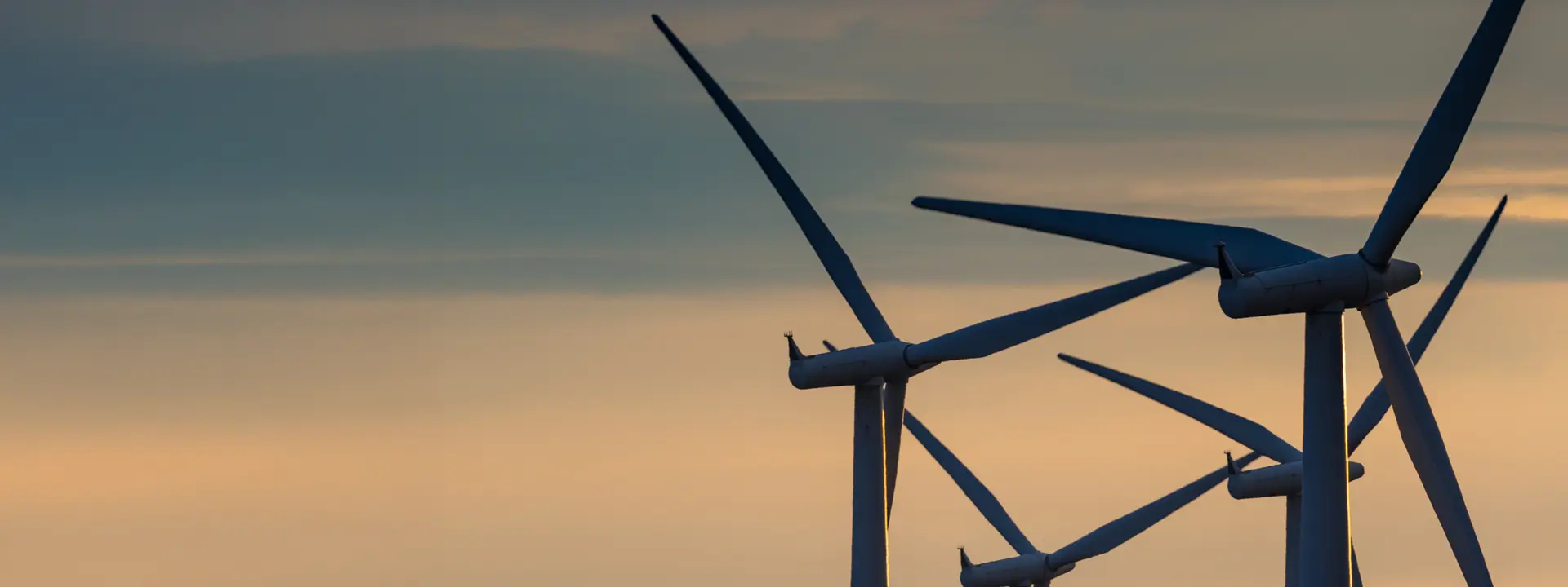 Silhouettes of wind turbines against a colorful sky during sunset, with soft clouds in the background.