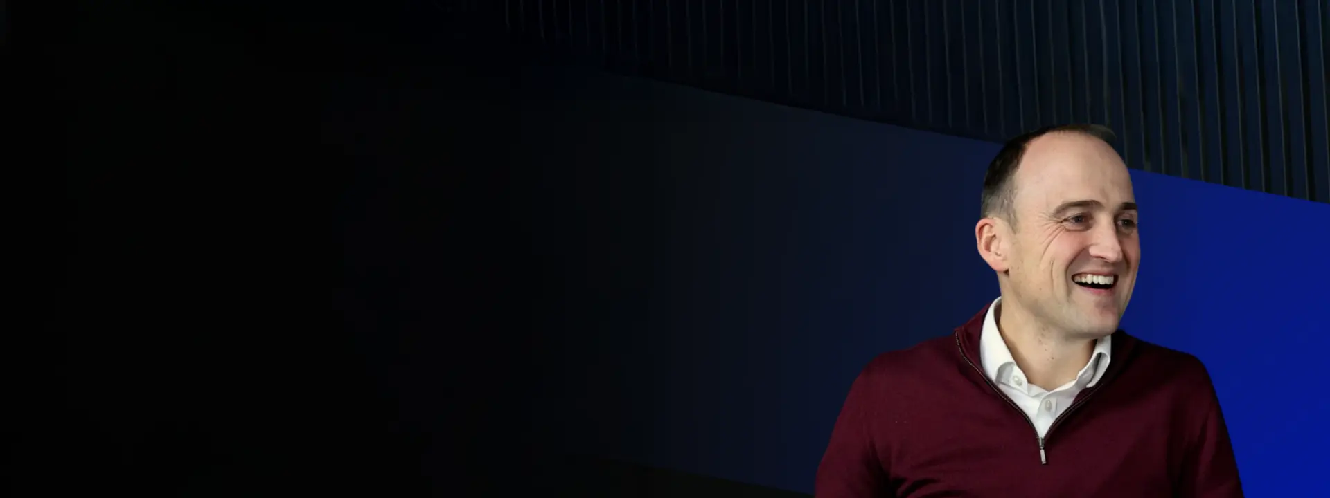 A man sitting and smiling, wearing a burgundy sweater and collared shirt, in front of a backdrop with dark and blue geometric shapes. The scene captures a relaxed and engaging atmosphere.