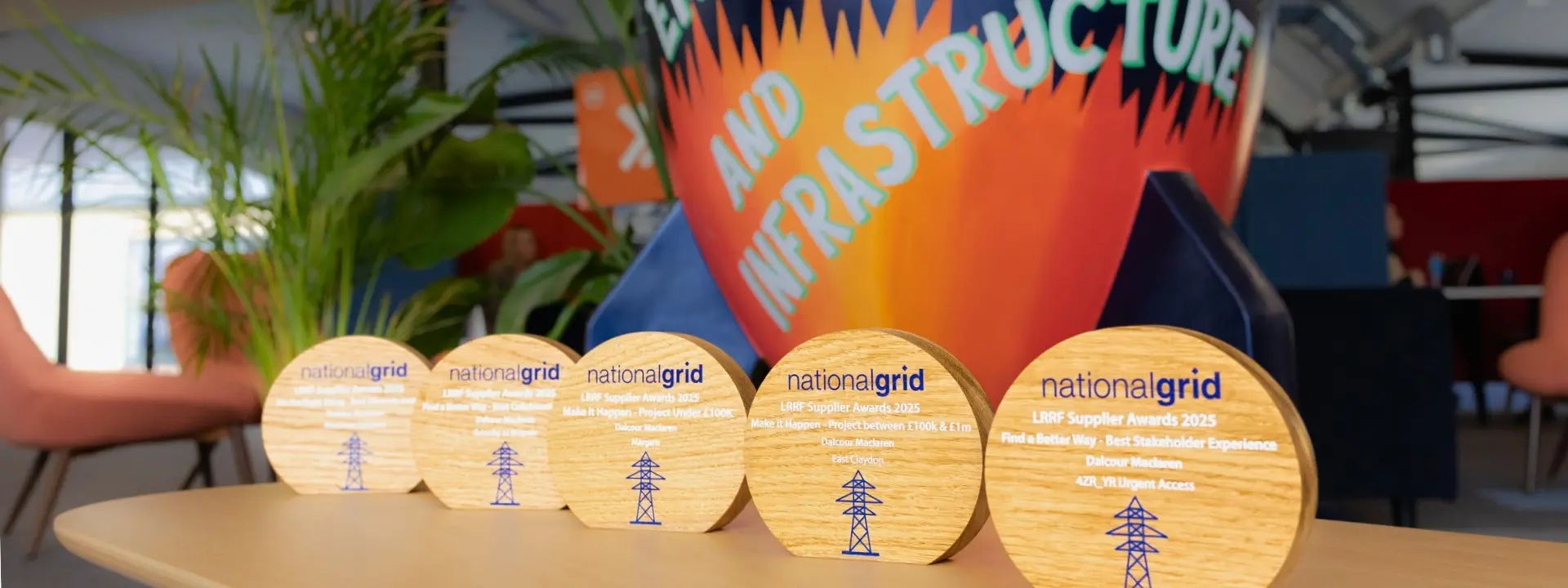 Five wooden awards with the text 'nationalgrid' and a design element depicting a power line, arranged on a table. In the background, a colorful banner that reads 'EMPOWERING COMMUNITIES AND INFRASTRUCTURE.'