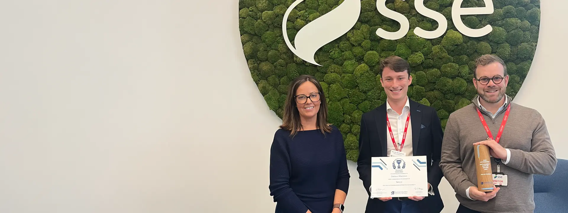 Three individuals standing in front of a moss-covered wall with the SSE logo. One person holds a certificate, while another holds a brown bag. They are posed in a modern office environment.