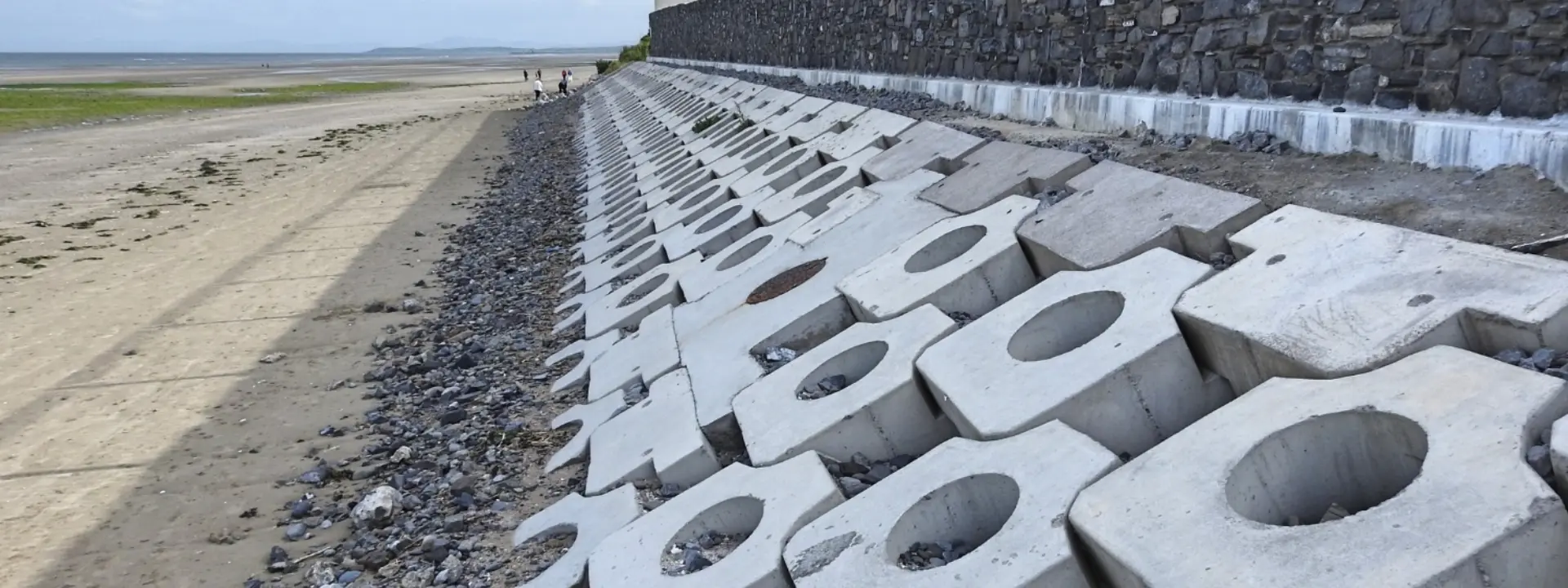 A series of large concrete blocks lined along a beach, with a stone wall in the background. The area is flat and dry, showing a mix of sand and pebbles, and a group of people can be seen walking along the shoreline.