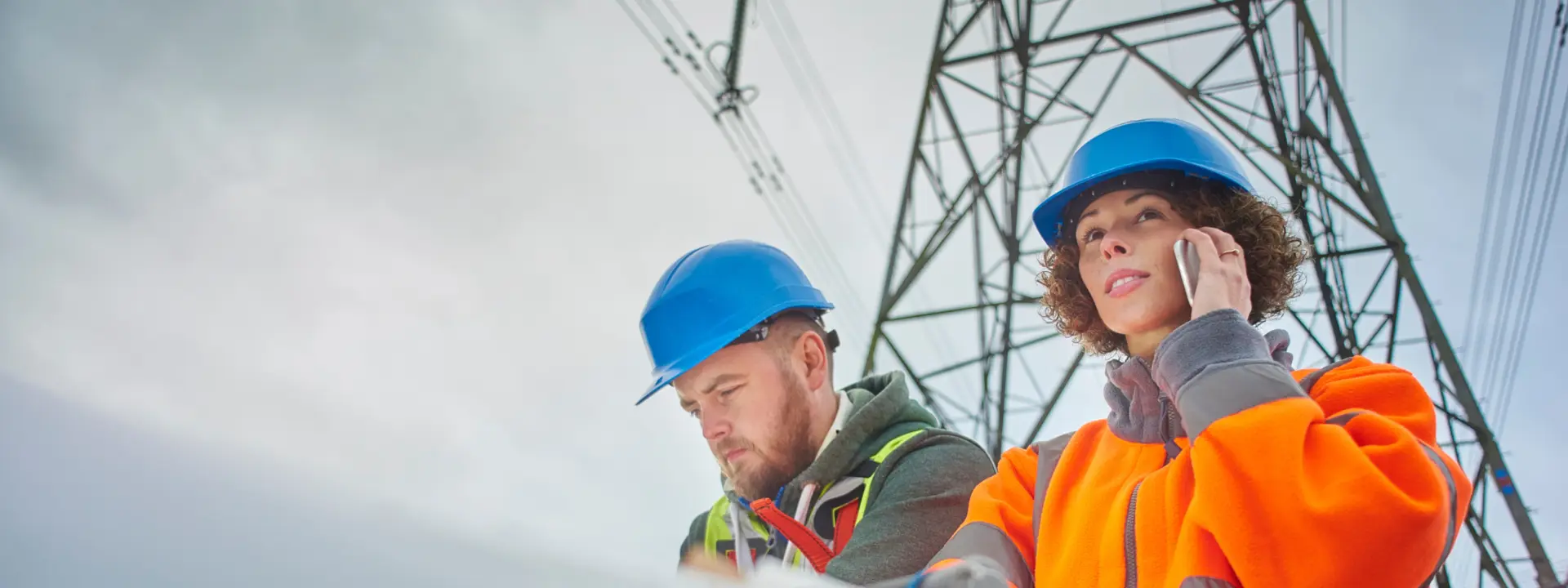 Two utility workers, one wearing an orange safety jacket and the other in a black and green outfit, are on-site near a power line tower. The woman is speaking on a phone while the man is focused on taking notes.