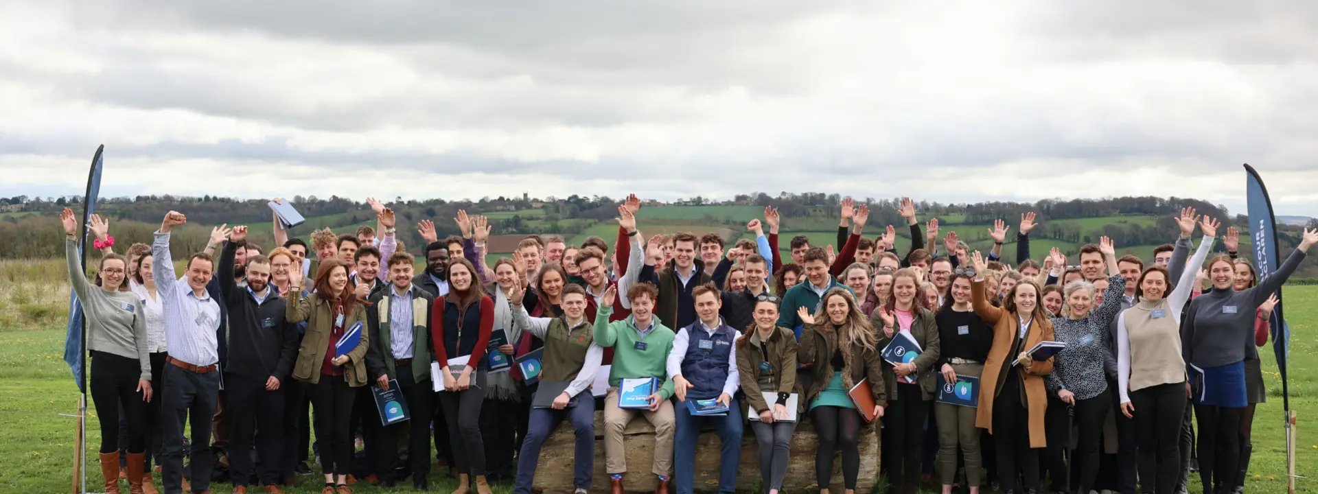 A large group of people, approximately 100, gathering outdoors with hands raised, smiling. They hold pamphlets and are standing on grass, with a cloudy sky and green fields in the background.
