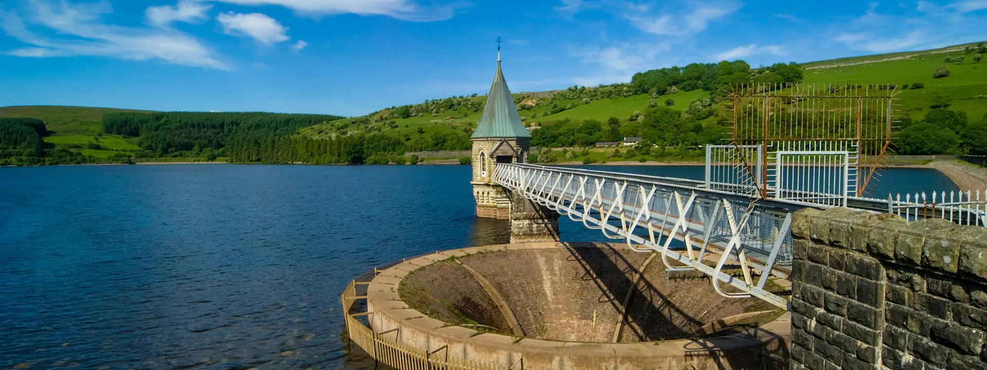 A scenic view of a reservoir with a stone tower and a bridge extending across the water. Lush green hills are visible in the background, while fluffy clouds float in a blue sky.
